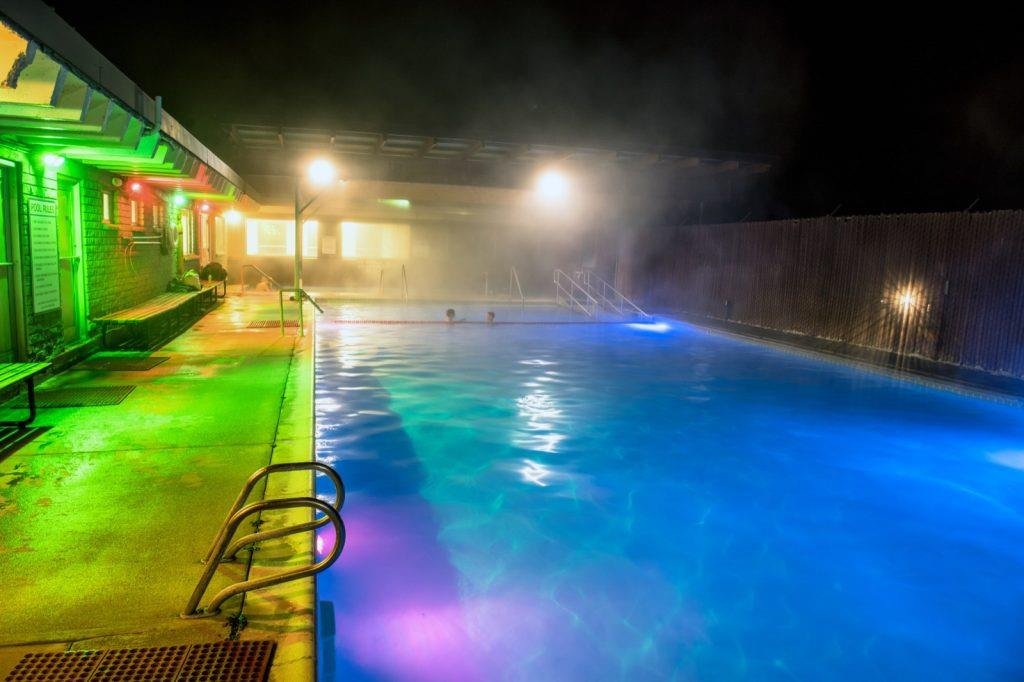 People relaxing in a steaming outdoor mineral pool at Heise Hot Springs surrounded by snow-covered ground.