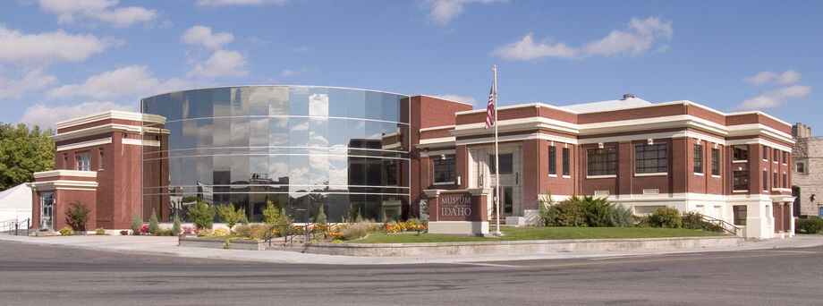 A wide exterior view of the Museum of Idaho in downtown Idaho Falls, featuring a mix of modern glass architecture and classic brickwork under a clear blue sky.