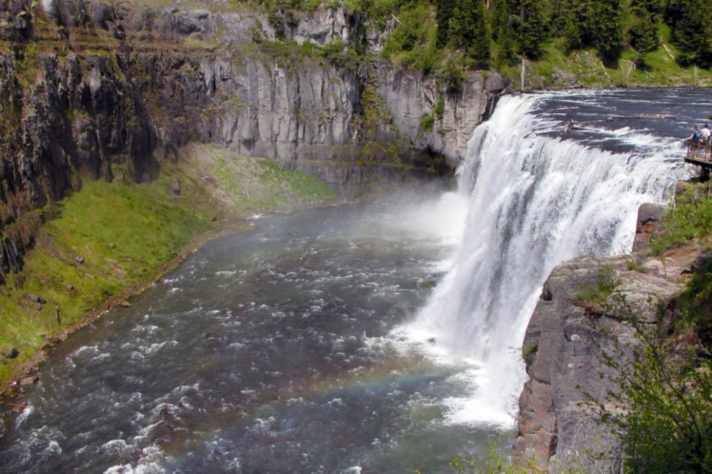 Powerful View of Mesa Falls A wide shot of a massive, thundering waterfall dropping into a deep canyon with mist rising from the bottom and thick pine forests on the surrounding cliffs.