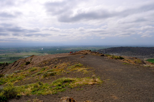 Hiking Trail at Menan Butte A dirt trail winding up the side of a volcanic cinder cone with green vegetation and a vast view of the Idaho plains under a cloudy sky.